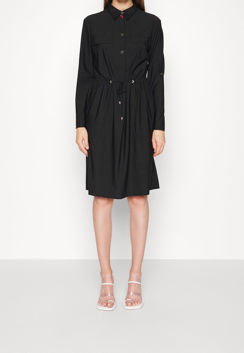 Femme portant une robe-chemise noire à manches longues longueur genou avec une taille à cordon et des sandales blanches à talons ouverts aux orteils.