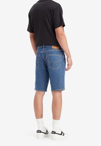 Man som bär blå denimshorts, svart t-shirt, vita strumpor och vita sneakers med svarta Levi's-logotypen, står med ryggen vänd mot betraktaren.
