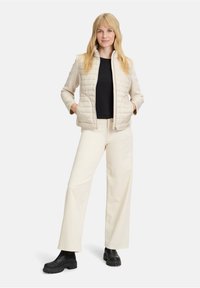 Beżowa puchowa kamizelka z kołnierzem typu stójka, zestawiona z czarną bluzką i kremowymi spodniami o szerokich nogawkach. Czarne botki za kostkę dopełniają stylizację.