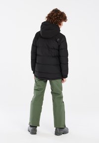 Doudoune noire avec capuche, présentant un design moderne et un motif matelassé horizontal, associée à un pantalon imperméable vert et des bottes grises.