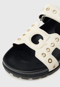 Λευκό σανδάλι από ψεύτικο δέρμα με χρυσούς κρίκους, ανοιχτή μύ τη και μαύρη υφασμάτινη σόλα. Διαθέτει χαρακτηριστικό στοιχείο με γράμμα κομμένο.