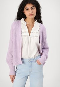 Lilla strikket cardigan med et teksturert mønster og knappedetalj, brukt over en hvit bluse med blonder og lyseblå jeans.