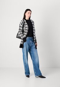 Cardigan preto e branco com letras abstratas, combinado com jeans de denim azul folgados e sapatos pretos de bico fino, apresentando uma mala preta.