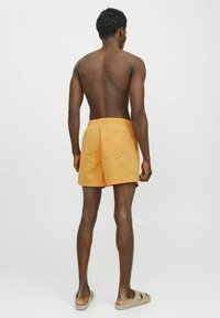 Shorts de bain orange pour homme avec une taille élastique et une poche arrière. Tissu lisse avec une coupe ample, portés avec des sandales beiges à enfiler.