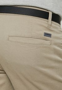 Beige bavlněné kalhoty s kapsou na zadní straně, doplněné o tmavý texturovaný pásek. Součástí je malá značka. Hladká textura, přiléhavý střih.