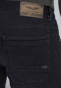 Jeans de mezclilla negra con una superficie texturizada, que presentan un diseño de bolsillo distintivo, etiqueta de cuero marrón y un sutil acento de logo bordado.