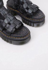 Sandalias de cuero negro trenzadas con suelas gruesas y texturizadas, y detalle de costura amarilla, reposando sobre una superficie clara.