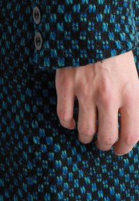 Il tessuto della giacca in misto lana presenta una base nera testurizzata con motivi a quadri blu e teal. Due bottoni scuri sono visibili sul polsino.