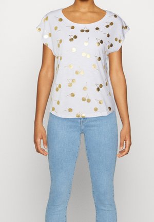 Vrouw draagt een witte top met korte mouwen met glanzende gouden kersenprints, gecombineerd met lichtblauwe skinny jeans.