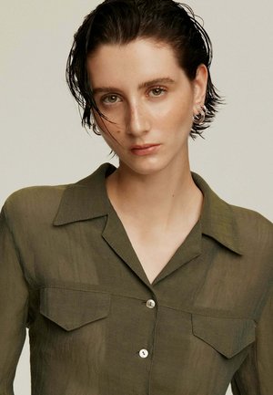 Camisa de botones verde oliva semitransparente con cuello, que presenta dos bolsillos delanteros y una textura sutil. El modelo posa con el cabello corto y con efecto mojado.