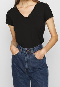Černý krátký rukáv top s výstřihem do v písmene V a krajkovými detaily na ramenou, spárovaný s modrými džíny vysokého pasu s předními kapsami a zapínáním na knoflík.