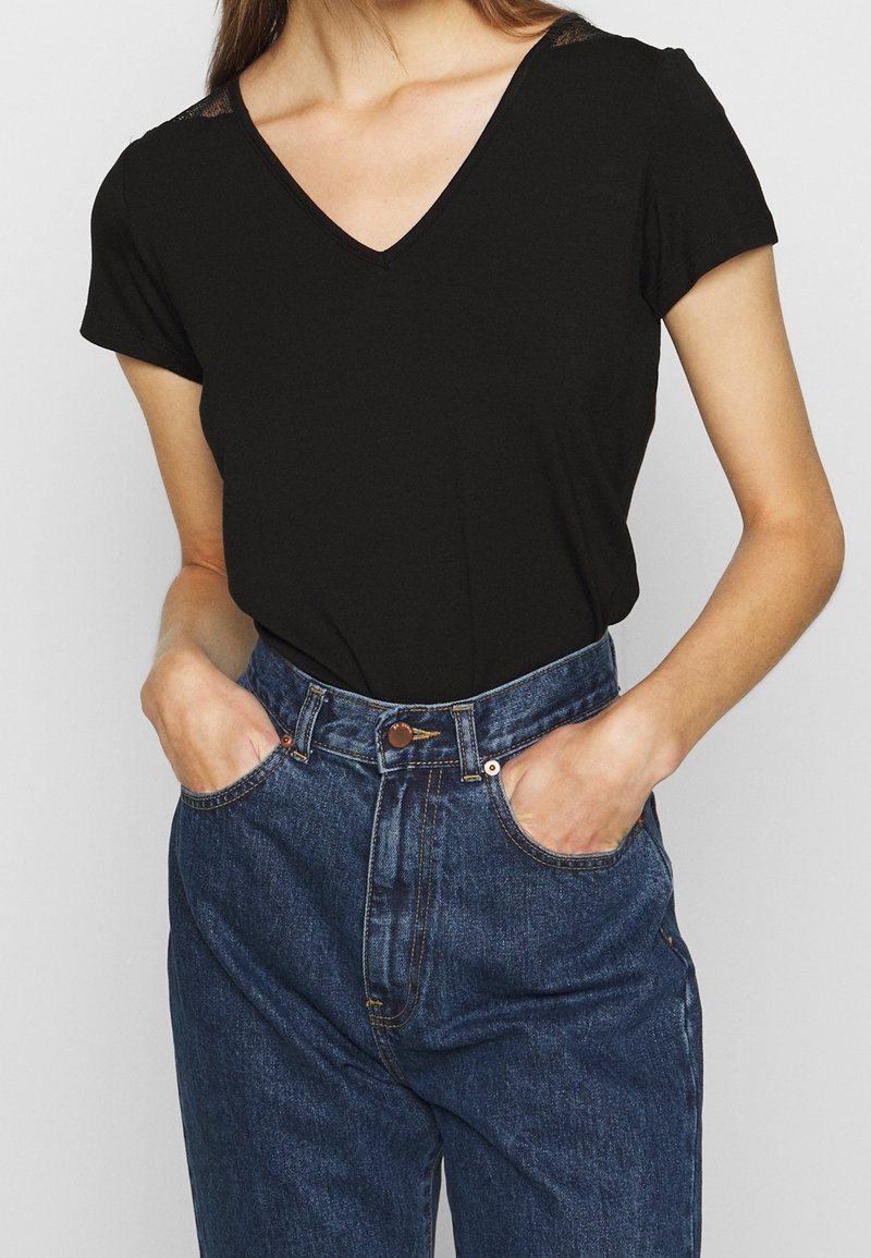 Černý krátký rukáv top s výstřihem do v písmene V a krajkovými detaily na ramenou, spárovaný s modrými džíny vysokého pasu s předními kapsami a zapínáním na knoflík.