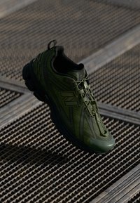 Zelená sportovní obuv s texturovaným síťovaným svrškem, zesílenou špičkou a vzorovanými akcenty. Obsahuje černou gumovou podrážku a šněrovací design.