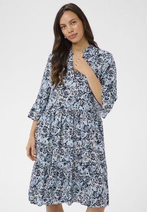 Hosszú sötét hajú nő térdig érő, laza, kék és fekete virágmintás ruhában, háromnegyedes ujjú, fehér háttéren.