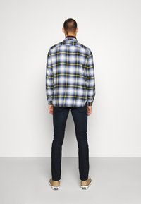 Camicia di flanella a quadri blu, bianca e gialla, con colletto classico e polsini abbottonati, abbinata a jeans scuri e scarpe beige.