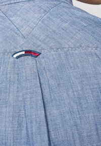 Camicia chambray azzurro chiaro con piega sul retro e un piccolo logo ricamato con accenti rossi, bianchi e blu. Tessuto morbido.