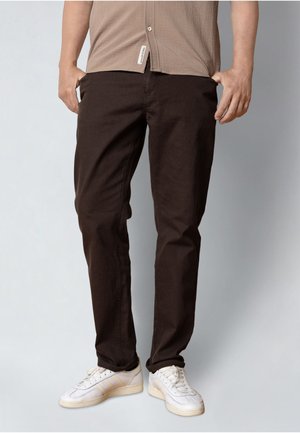 Mann trägt dunkelbraune Hose, beiges, strukturiertes Hemd und weiße Turnschuhe, steht mit den Händen in den Taschen vor einem neutralen Hintergrund.