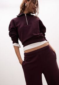Felpa bordeaux con colletto con zip, abbinata a pantaloni felpati dalla vestibilità ampia. La felpa presenta dettagli a coste, creando una texture.