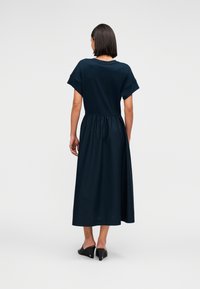 Φόρεμα μήκους μέχρι το γόνατο σε ναυτικό μπλε, με κοντά μανίκια, ζώνη στη μέση και λεία υφή υφάσματος. Φορεμένο με μαύρες ανοιχτές γόβες.