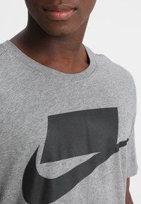 Šedé bavlněné tričko s kulatým výstřihem. Na přední straně velký černý grafický motiv připomínající logo Nike. Krátké rukávy a měkká textura.