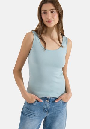 Femme aux longs cheveux bruns portant un haut sans manches bleu clair et un jean bleu, debout les mains dans les poches sur fond blanc.
