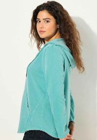 Vihreä huppari, jossa on kengurupocket, kontrastiväriset saumat ja pehmeä, tekstuurinen huppu. Silkkinen kangas, jossa on rento istuvuus.