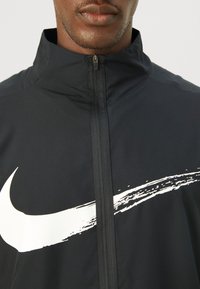 Sort zip-jakke lavet af glat stof med høj krave. Har et stort hvidt Nike-logo og en tekstureret lynlåsdetalje.