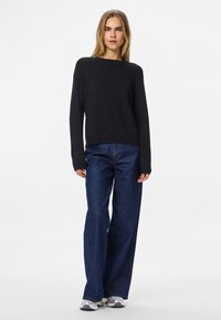 Nő áll, fekete hosszú ujjú pulóvert, szélvastag kék farmert és fehér sportcipőt viselve, egy sima fehér háttér előtt.