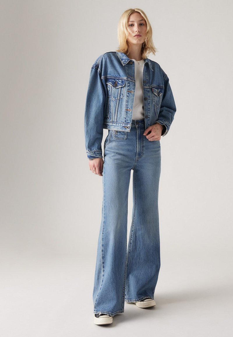 Veste en jean avec une coupe courte, de couleur bleu clair, et fermeture à boutons. Jean léger à jambes larges avec une texture délavée. Chemise blanche en dessous.