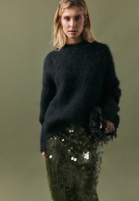 Black fuzzy sweater with a crew neckline, paired with a green sequined skirt. Black fringed handbag adds texture. Background is muted green.