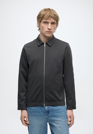 Ung mand med blondt hår iført en mørkegrå lynlåstrøje og lyseblå jeans, stående mod en ensfarvet hvid baggrund.