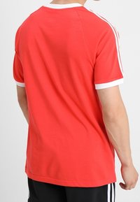 Punainen puuvillainen T-paita valkoisella reunuksella, lyhyet hihat ja kolme valkoista raitaa olkapäiden kohdalla. Suora leikkaus, yksivärinen malli.