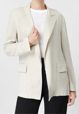 Person trägt einen hellbeigen Blazer über einem weißen T-Shirt und schwarzen Hosen, mit einer Hand, die das Revers des Blazers anpasst.