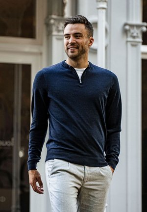 Marineblauwe trui met een halve rits halslijn, gecombineerd met lichtgrijze geblokte broek. De stof lijkt zacht met een lichte textuur.