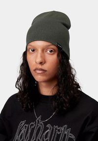Gorro de punto color verde oliva con un ajuste holgado. Cuenta con una etiqueta en el lateral. Se lleva con una sudadera negra que muestra letras blancas.