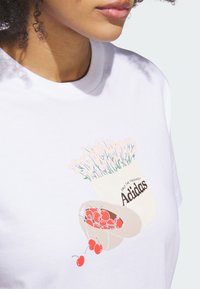 Biela bavlnená tričko s grafickým dizajnom čerešní a kvetináča, s textom "Adidas" v čiernej farbe, vytvárajúcim hravú vizuálnu tému.