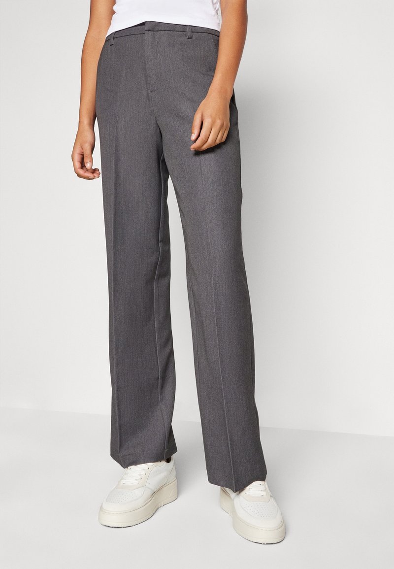 Personne portant un pantalon gris droit et des baskets blanches à plateforme, debout contre un fond clair uni.