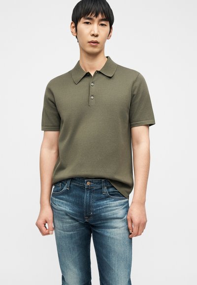 Νέος άνδρας που φοράει κοντομάνικο πόλο σε ελιά πράσινο και ξεθωριασμένο μπλε τζιν, στέκεται μπροστά σε ένα απλό λευκό φόντο.