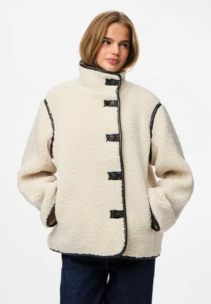 Vrouw in een crèmekleurige sherpa button-up jas met zwarte afwerking, staand met handen in de zakken en iets naar de zijkant kijkend.