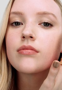 Close-up van een gezicht met een gladde huid, lichte sproeten en een neutrale lipkleur. Het onderwerp brengt make-up aan met een hand versierd met een ring.