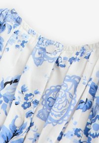 Balta medžiaga su mėlynu gėlių ir nėrinių raštu, puošta skaidriomis apvalios formos sagutėmis ir raukiniais ties iškirpte.