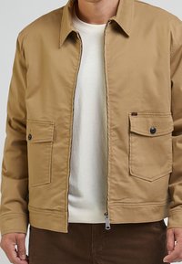 Beige Baumwolljacke mit Reißverschluss, zwei Brusttaschen und einem strukturierten Kragen. Sie verfügt über eine glatte Textur und ein minimalistisches Design.
