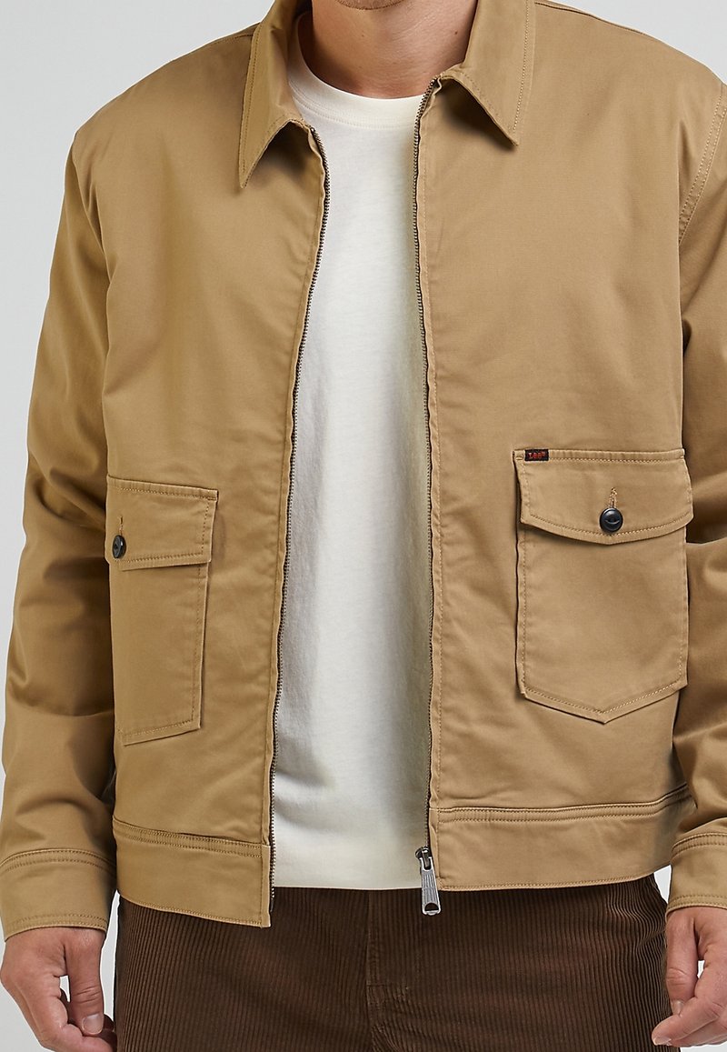 Beige Baumwolljacke mit Reißverschluss, zwei Brusttaschen und einem strukturierten Kragen. Sie verfügt über eine glatte Textur und ein minimalistisches Design.