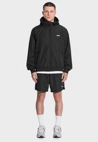 Czarna kurtka z kapturem, na suwak z przodu i elastycznymi mankietami; w połączeniu z czarnymi szortami. Materiał wydaje się lekki i wodoodporny.