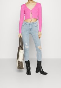 Rosa ribbad kort cardigan med knyt fram, matchad med ljusblå trasiga skinny jeans och svarta klackiga ankelskor, hållande en brun och vit tote-väska.