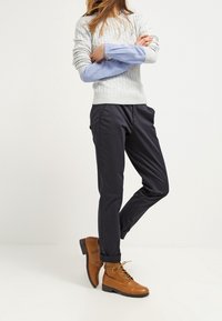 Maglione grigio chiaro a maglia grossa, camicia blu a fantasia sotto, pantaloni blu scuro e stivali alla caviglia marroni con lacci.