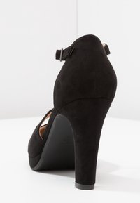 Chaussure à talon haut en daim noir avec un bout arrondi et une bride à la cheville, dotée d'un talon robuste et de détails découpés sur les côtés.