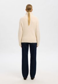 Grietos spalvos laisvo silueto megztinis su aukštu apykakliu ir raštuotu apačia, derinamas su tamsiais džinsais. Lygus paviršius ir laisva forma.