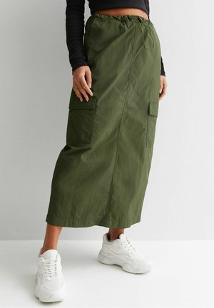 Personne portant une longue jupe cargo vert olive avec des poches et une taille avec cordon, associée à des baskets blanches épaisses.