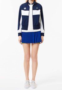 Veste zippée bleu marine et blanche avec col côtelé ; jupe plissée bleue ; baskets blanches. Accents de logo notables sur la veste.